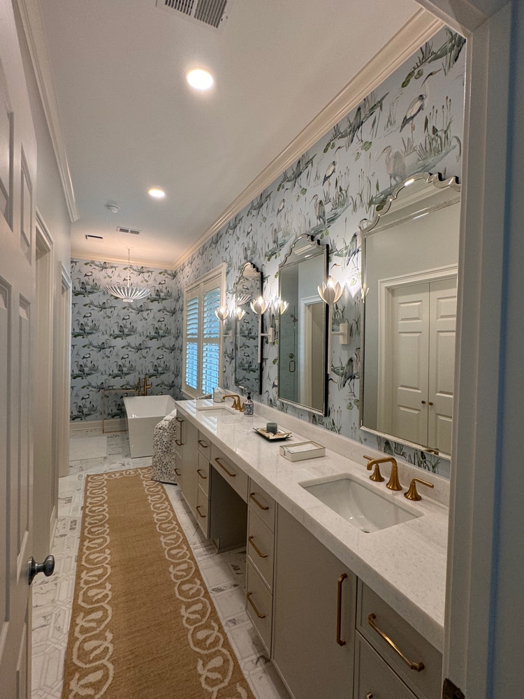 Dual-sink bathroom with white quartz counters, gold fixtures, and large mirrors, botanical wallpaper, and a freestanding tub at the far end.